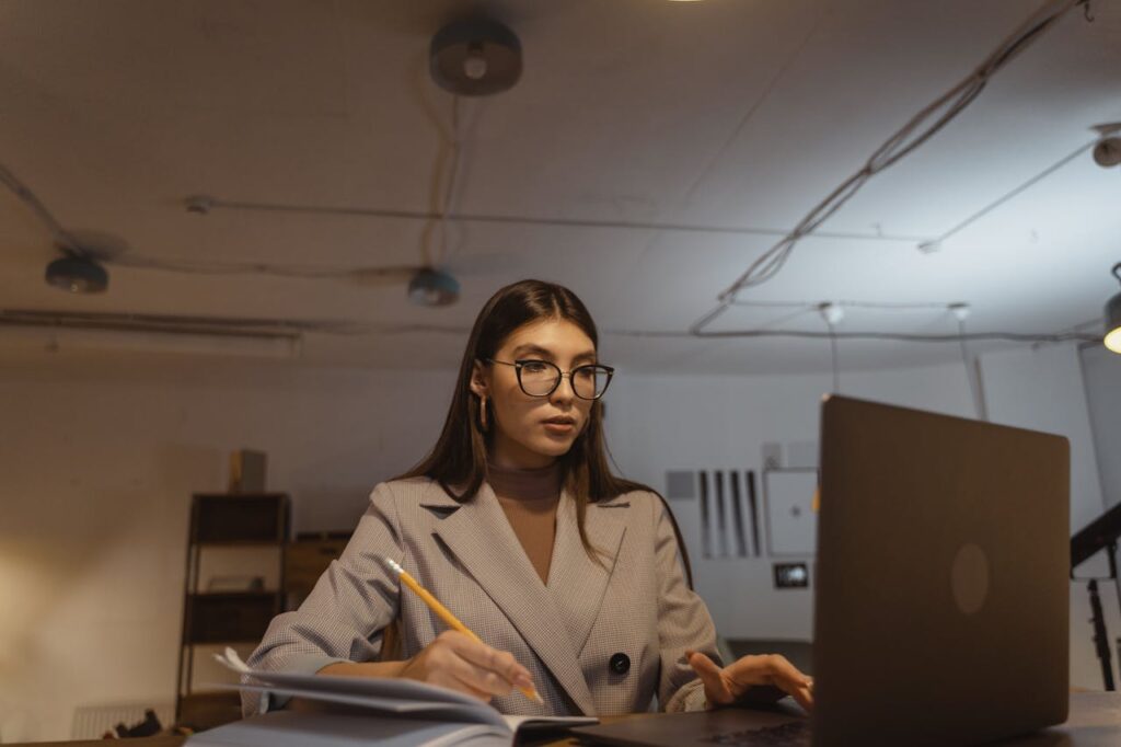 Focused young businesswoman in glasses working late on a laptop in a modern office setting, taking notes.